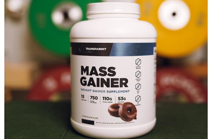 Transparent Labs Mass Gainer in a white container with black and blue writing, blurred weight plates in the background